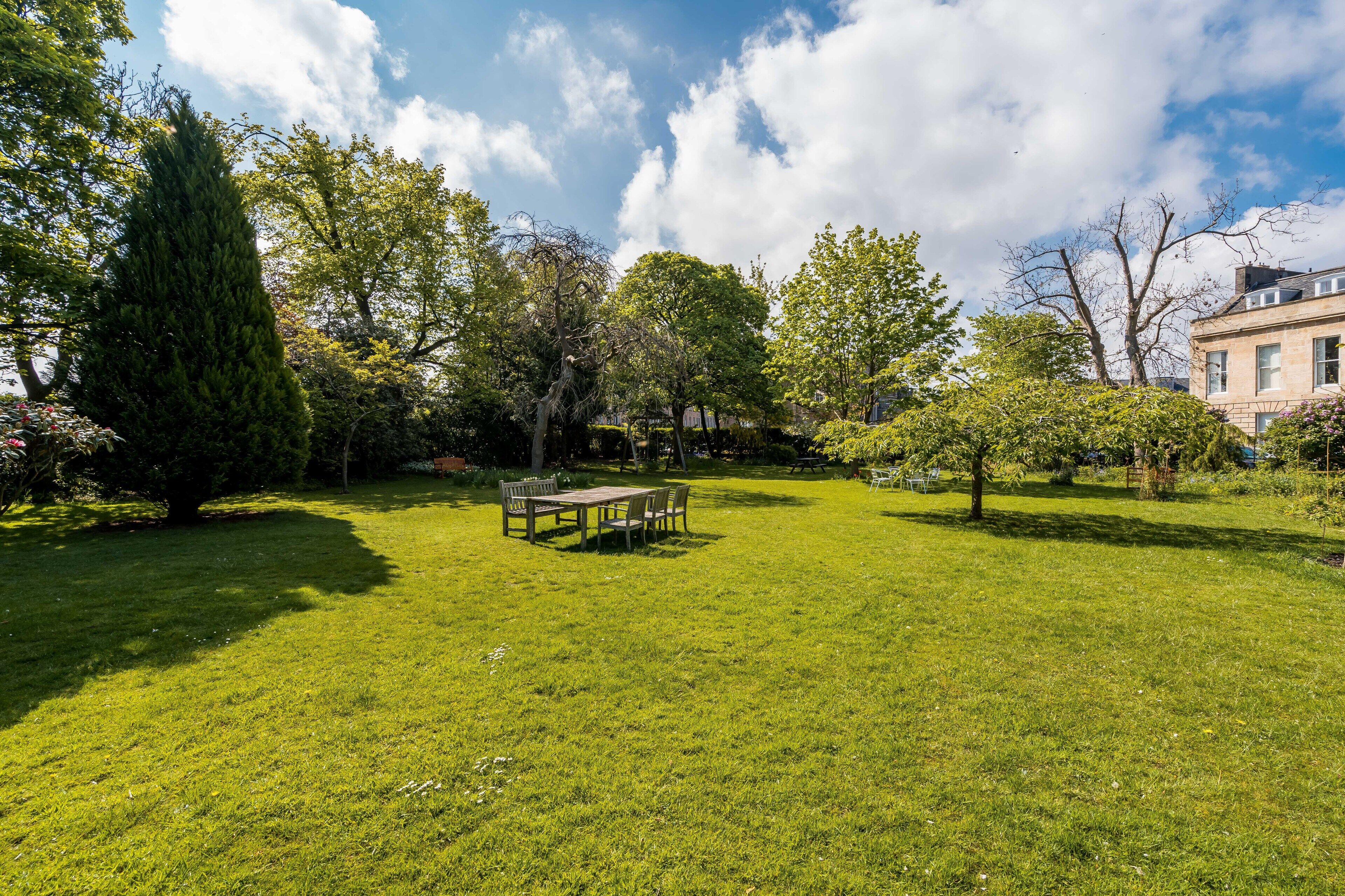 A well-maintained garden with a wooden table and chairs, surrounded by various trees and shrubs.
