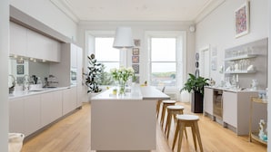 A modern kitchen with a central island, wooden stools, and a view through large windows. - Crescent House (Edinburgh)