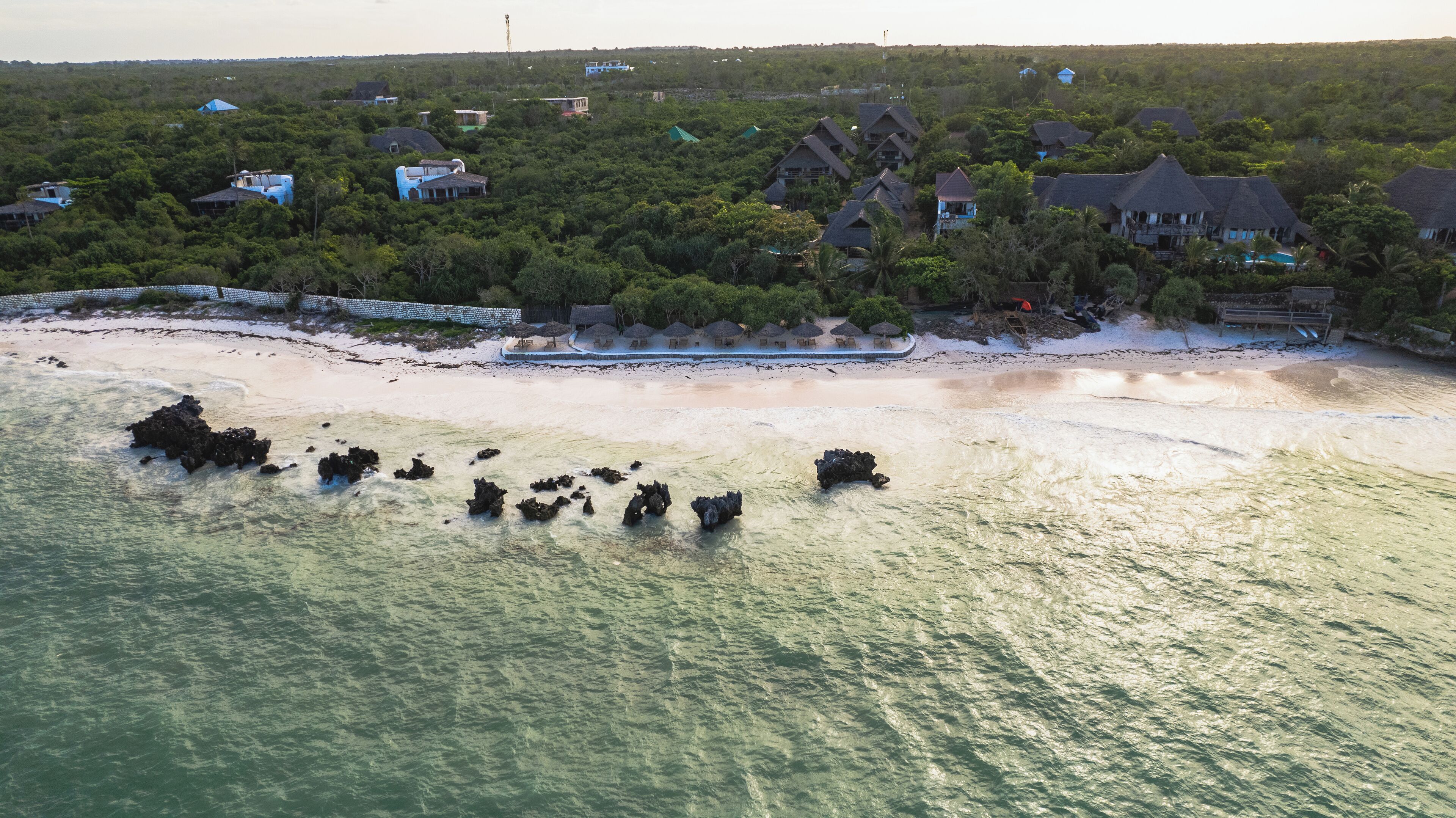 An aerial view of a coastal area with houses surrounded by dense vegetation, a sandy beach, and rocky outcrops in the water.