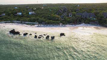 An aerial view of a coastal area with houses surrounded by dense vegetation, a sandy beach, and rocky outcrops in the water.