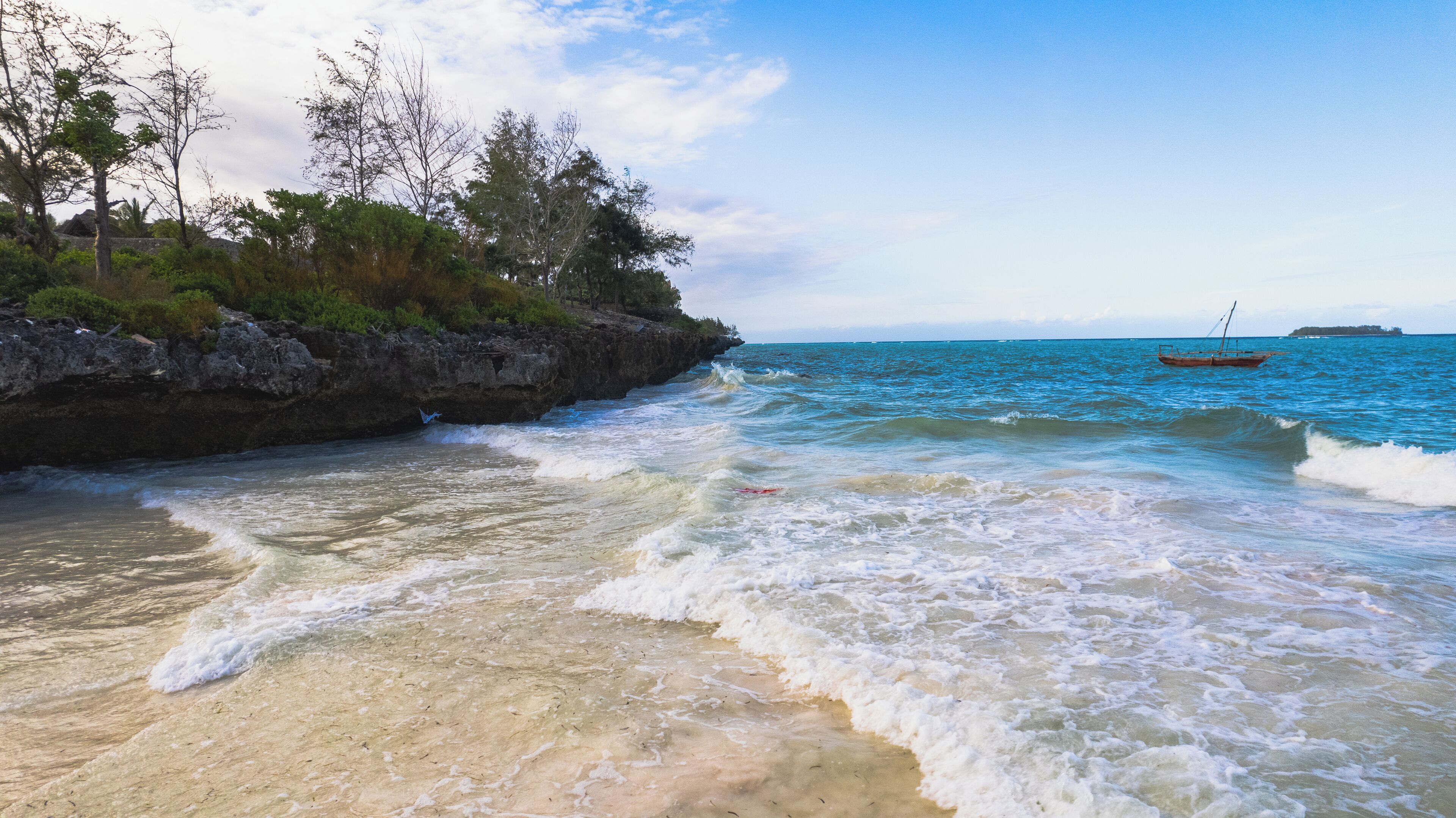 A beach with clear waters, a rocky shoreline, and a boat in the distance.
