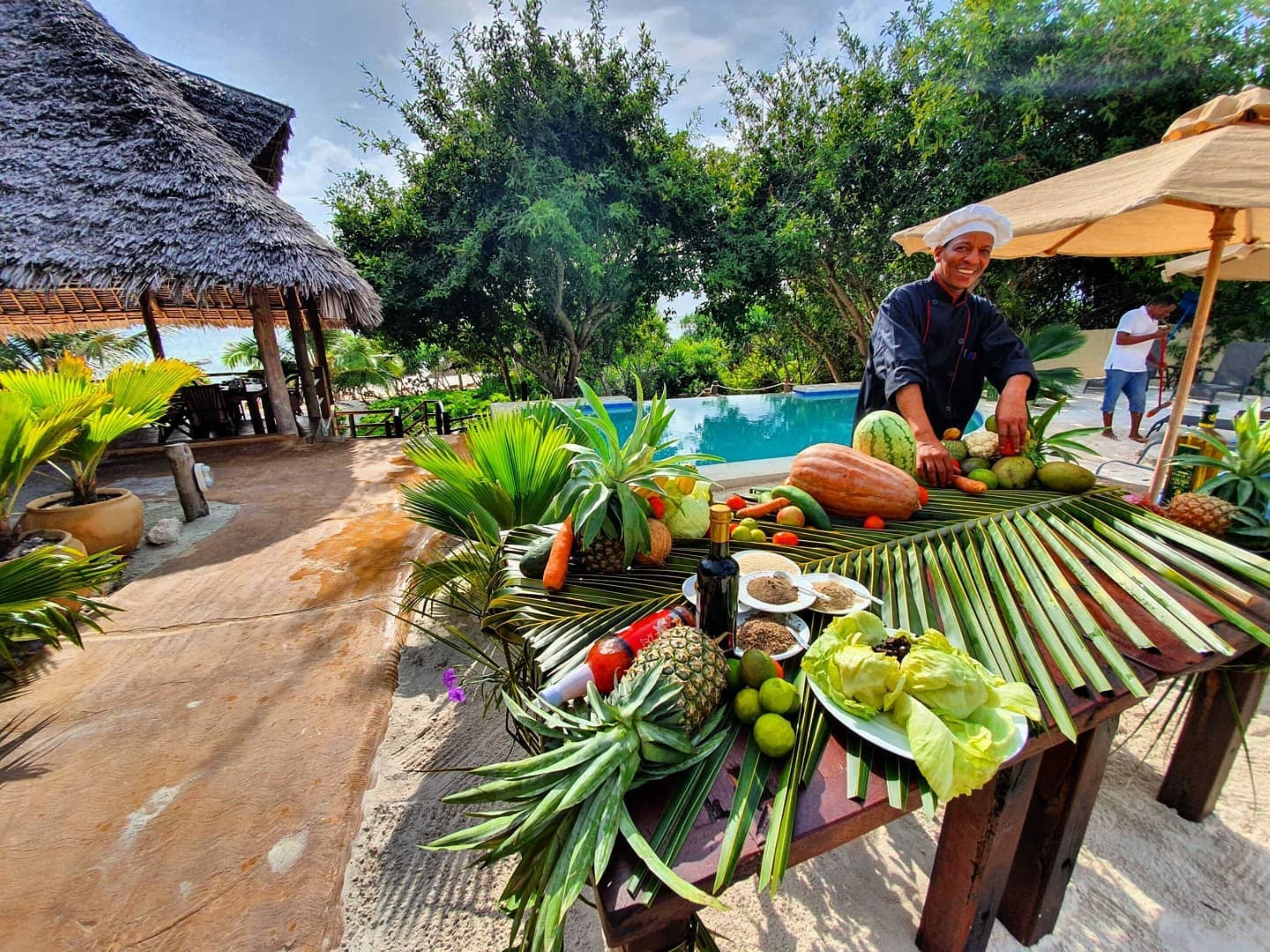 A tropical outdoor setting with a table displaying various fruits and vegetables, a pool in the background, and a thatched-roof structure.