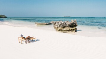 Una playa con arena blanca, una gran formación rocosa y una silla de madera con sombrero y libro.
