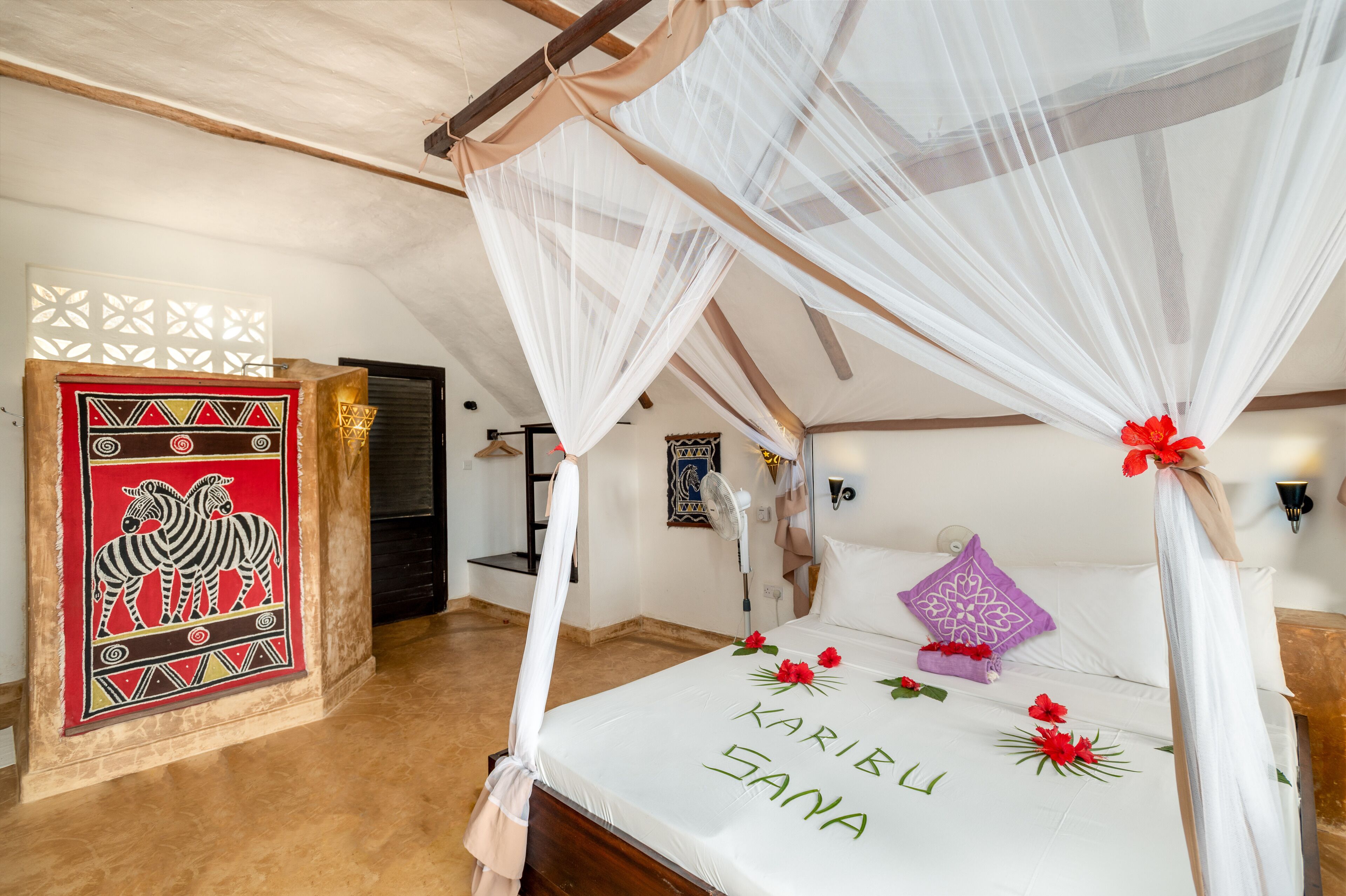 A room with a bed under a canopy, a zebra-themed artwork on the wall, and a bed with 'Karibu Sana' written on it.