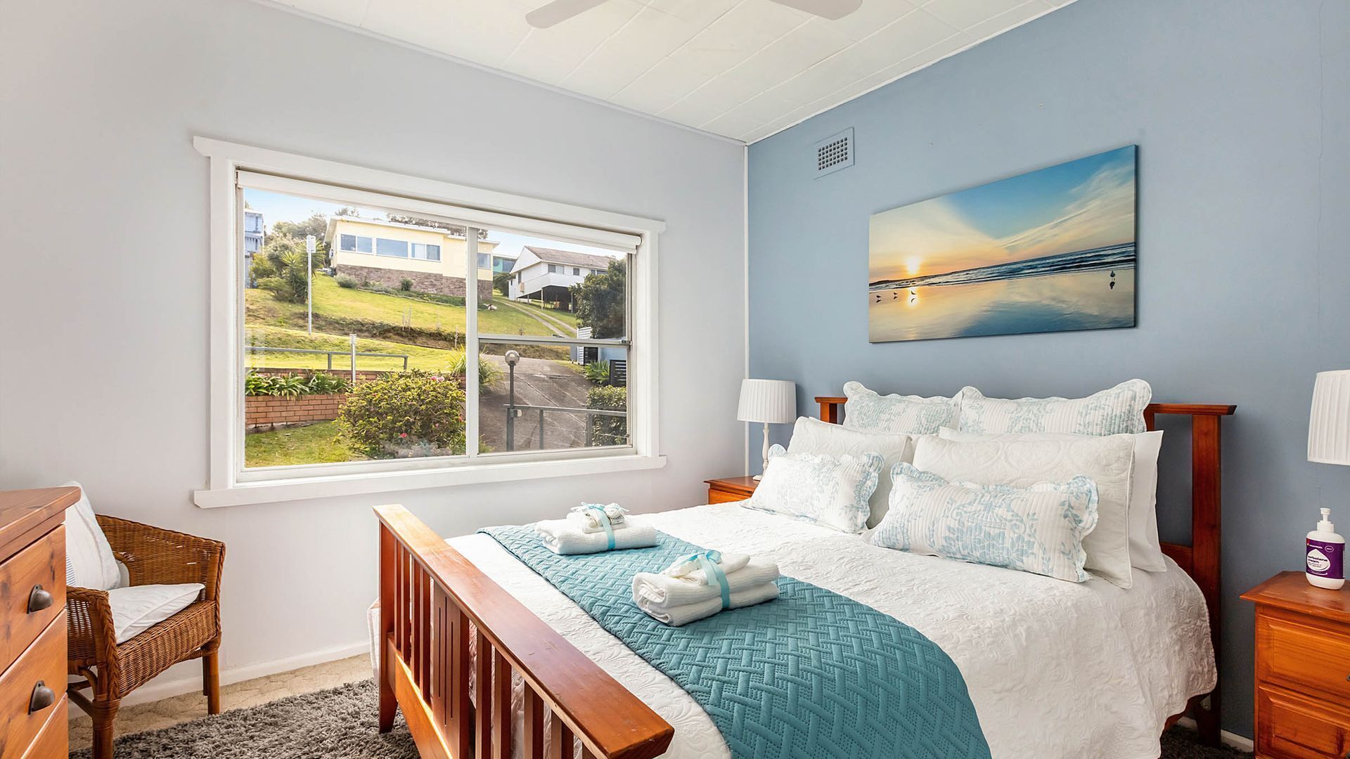 A bedroom with a large window, a bed with a blue quilt, a painting of a sunset, and a view of houses outside.