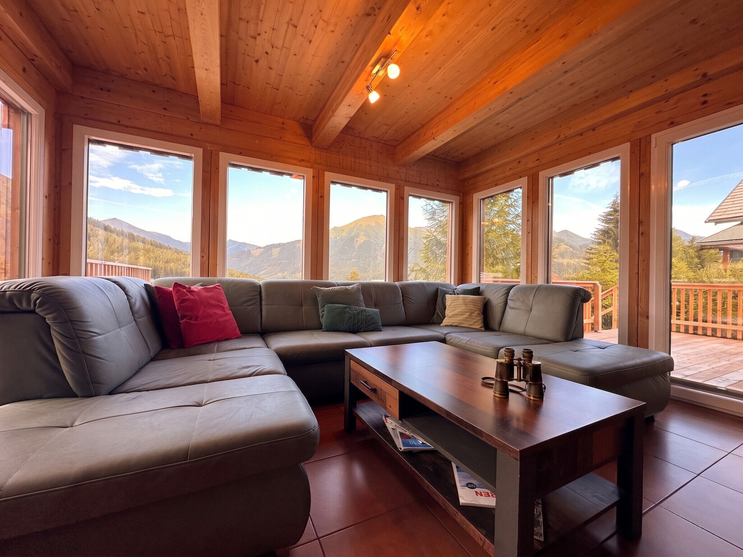 Un intérieur moderne avec un canapé d’angle, une table basse en bois et de grandes fenêtres offrant une vue sur les montagnes.