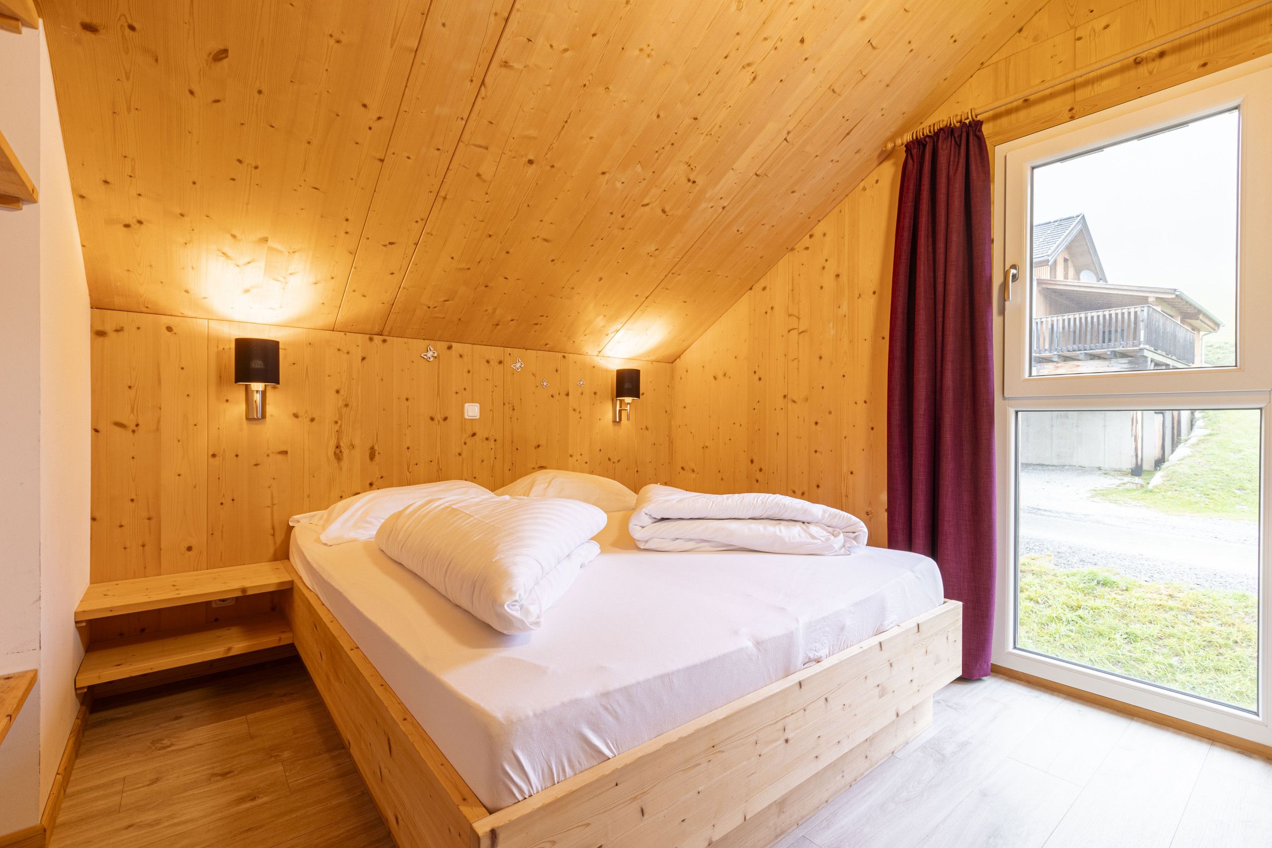 Une chambre dans une cabane en bois, avec un lit, deux lampes fixées au mur, une fenêtre avec des rideaux et une vue sur un bâtiment à l’extérieur.