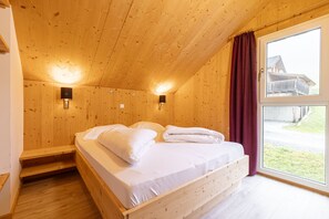 A wooden cabin bedroom with a bed, two wall-mounted lights, a window with curtains, and a view of a building outside. - Superior Chalet # 61 with IR-Sauna (Hohentauern)