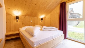 A wooden cabin bedroom with a bed, two wall-mounted lights, a window with curtains, and a view of a building outside. - Superior Chalet # 61 with IR-Sauna (Hohentauern)