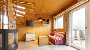 A cozy room with a bed, a small wooden table, a TV mounted on the wall, and a large window with a view of a wooden deck. - Superior Chalet # 61 with IR-Sauna (Hohentauern)