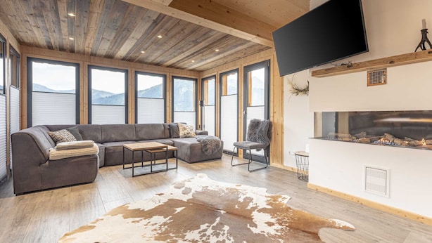 Un soggiorno moderno con un ampio divano componibile, una TV a schermo piatto montata a parete, un caminetto e ampie finestre con vista sulle montagne.