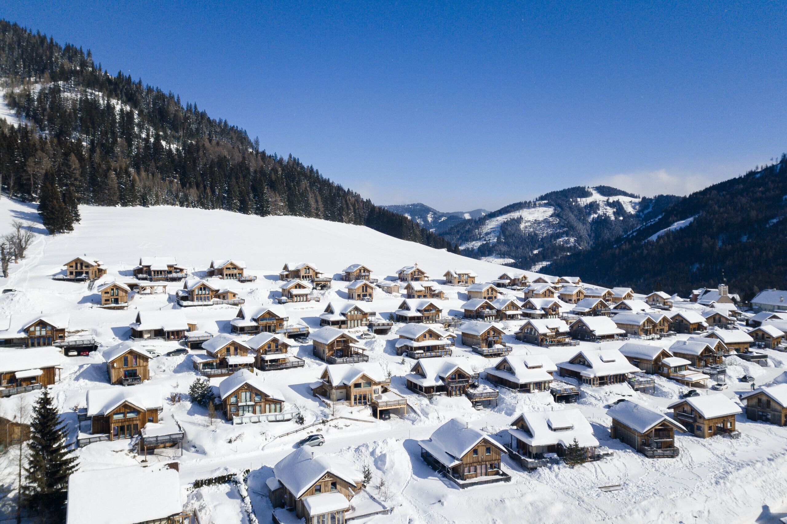 A snow-covered village with wooden cabins, surrounded by mountains and trees.