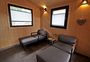 A room with two grey sofas, a small table with a plant, and two windows with blinds.