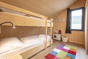 A wooden cabin room with bunk beds, a window with a view, a small kitchenette, and a colorful rug.