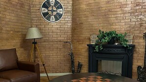 A room with a brown leather sofa, a round table with a checkered top, a clock on the wall, a floor lamp, a fireplace with a plant, and a saxophone against the wall. - Bamboo Boutique (Lilongwe)