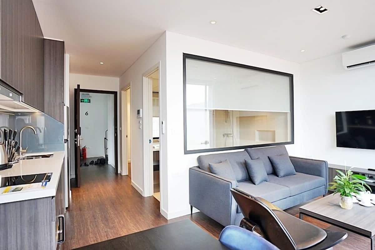 A modern hotel room with a kitchenette, a grey sofa, a flat-screen TV, and a large mirror.