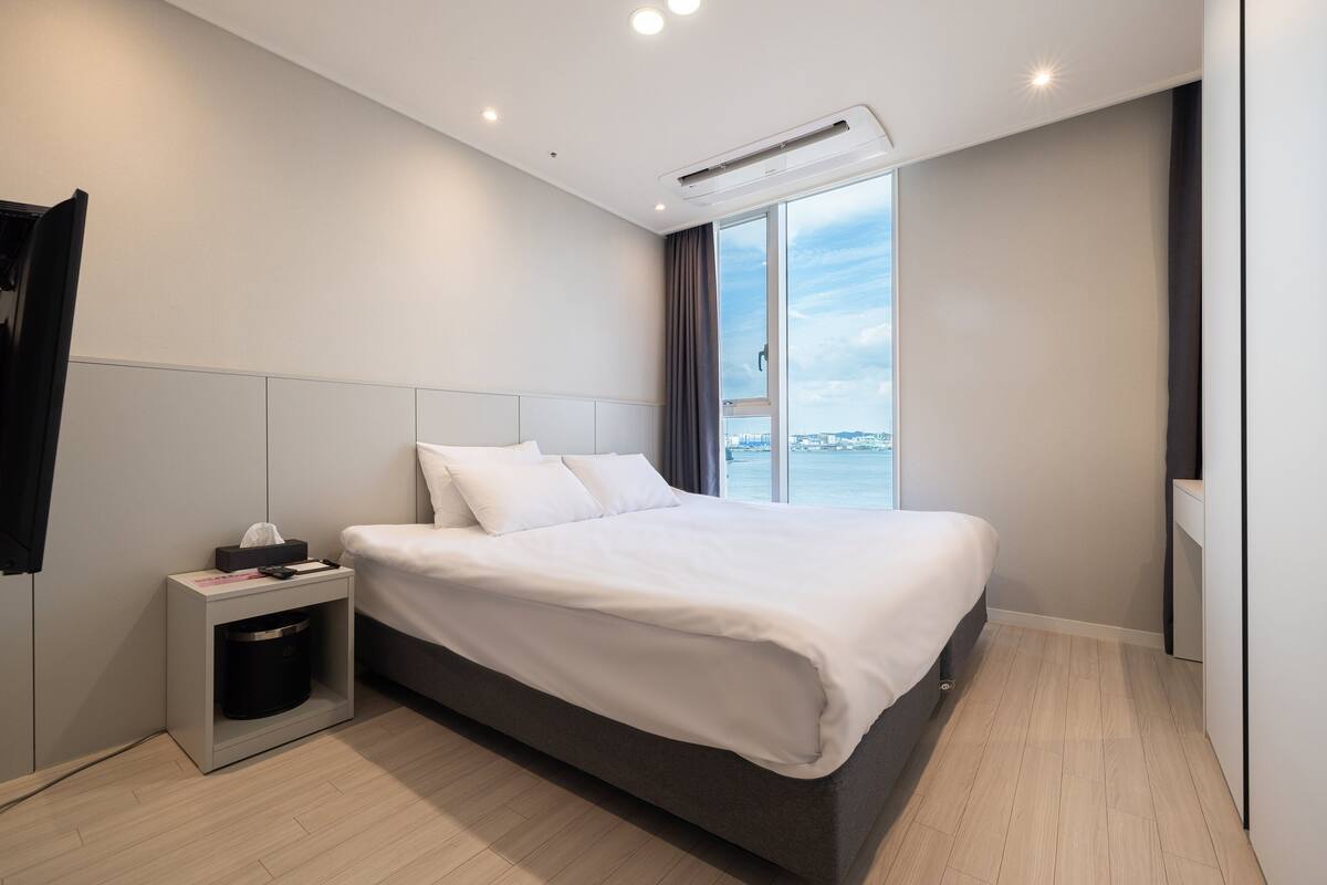 A modern hotel room with a large bed, a bedside table, a built-in heater, and a view of the sea through a large window.