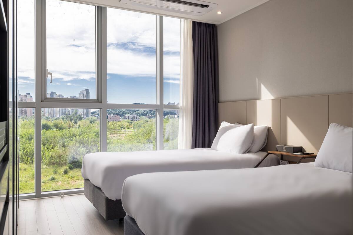 A hotel room with two beds, a large window with a city view, a bedside table with a lamp, and a wall-mounted TV.