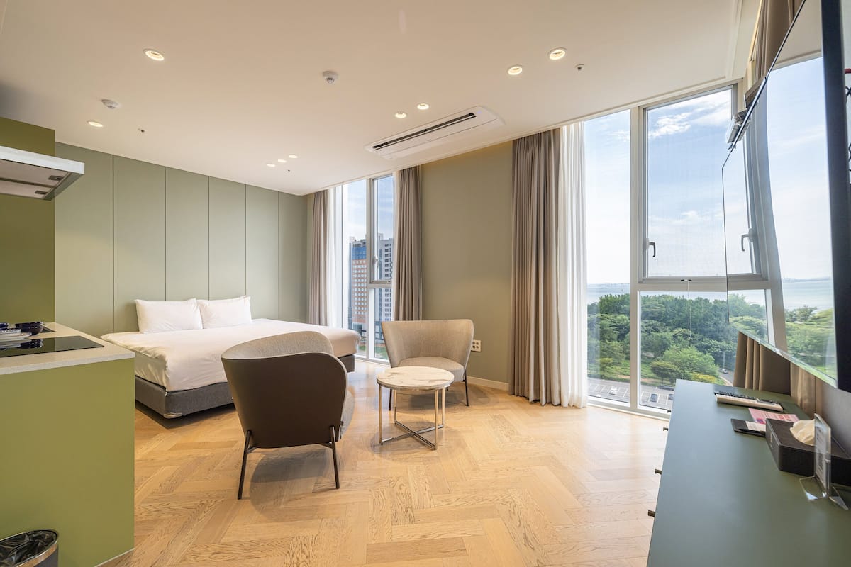 A modern hotel room with a bed, two chairs, a desk, and large windows offering a city view.