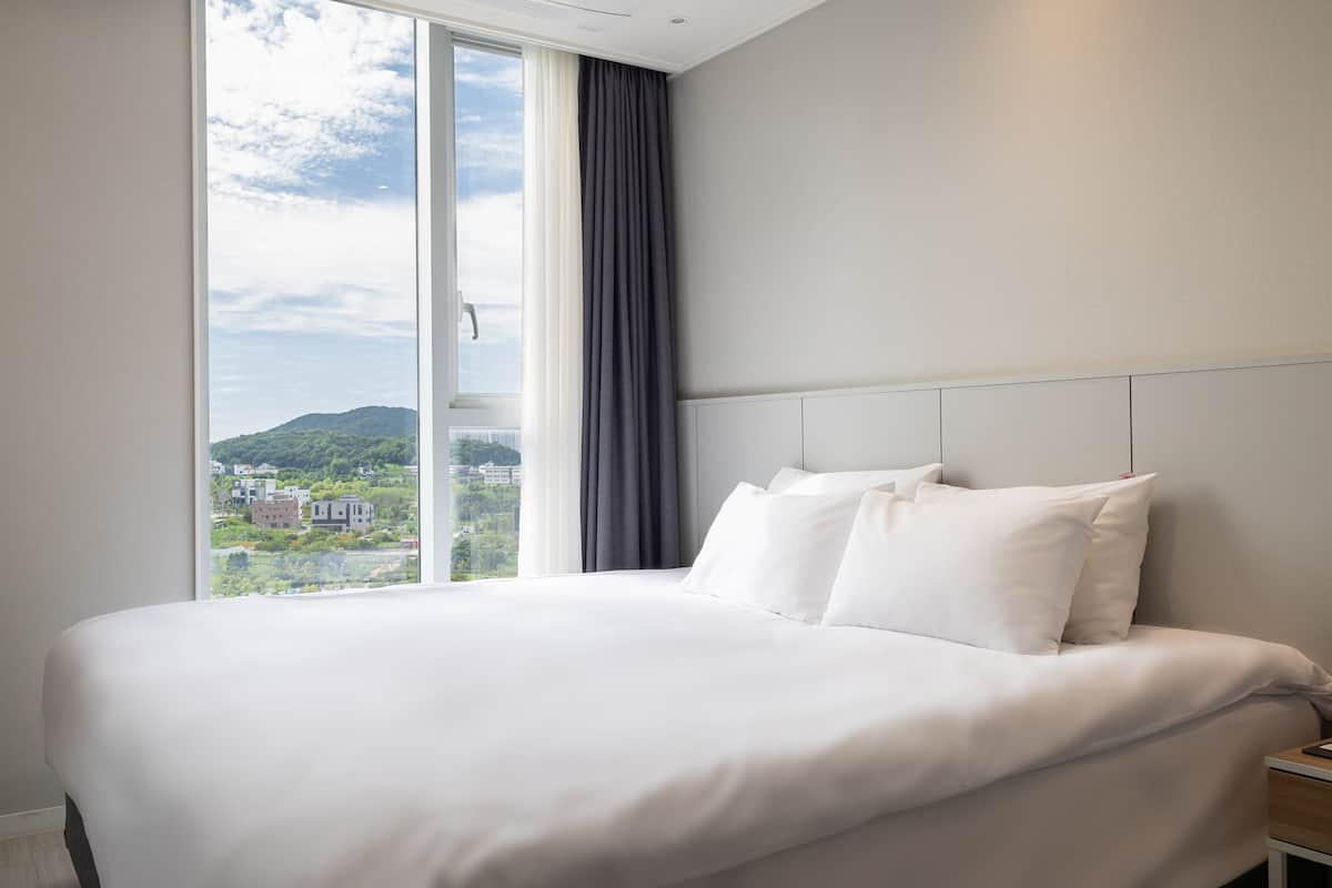 A hotel room with a large bed, a nightstand, and a view of a cityscape through a window.