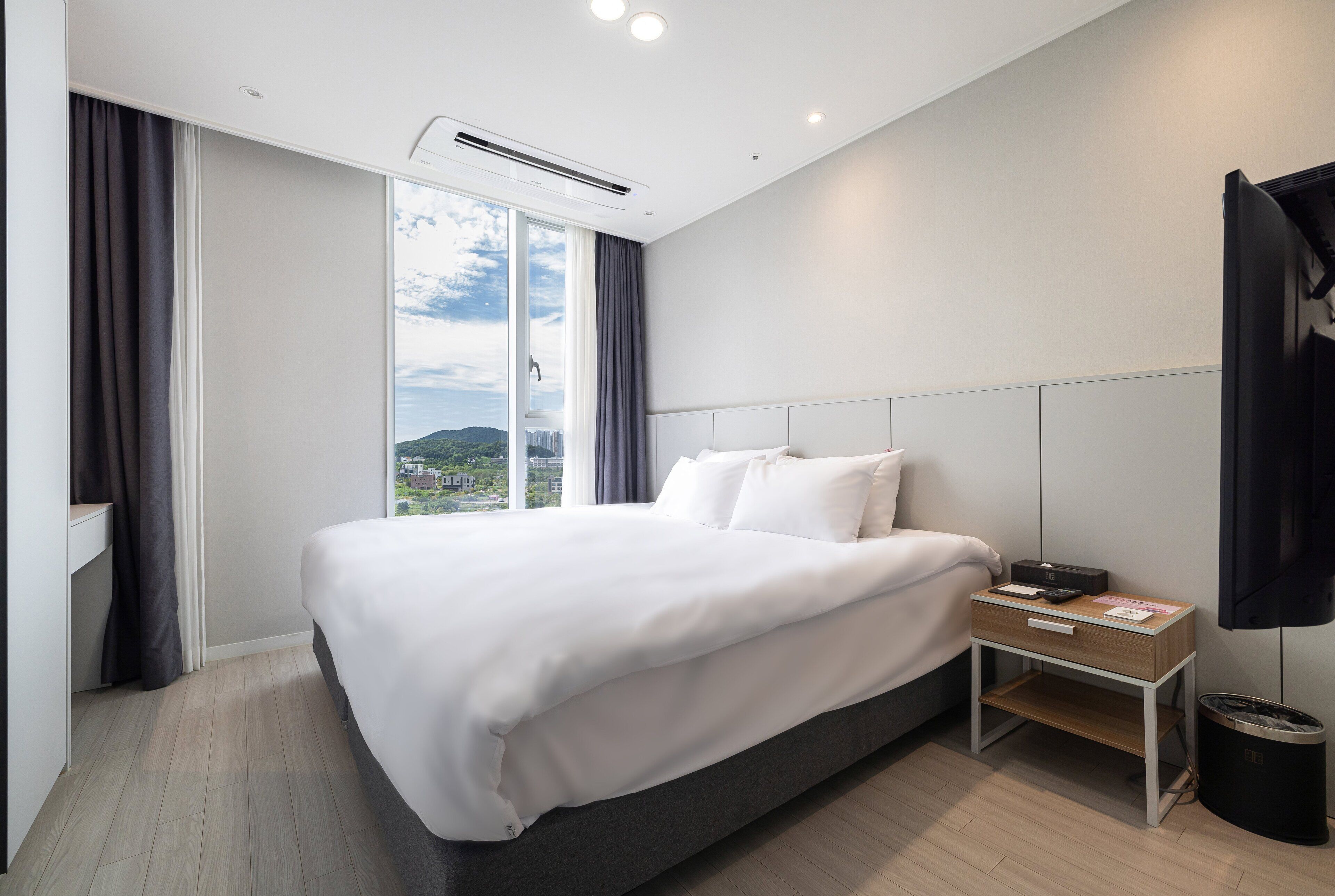 Une chambre d’hôtel moderne avec un grand lit, une table de chevet avec une lampe, une vue sur le paysage urbain à travers une grande fenêtre et une poubelle noire.