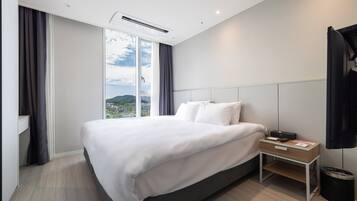 A modern hotel room with a large bed, a bedside table with a lamp, a view of a cityscape through a large window, and a black trash bin.