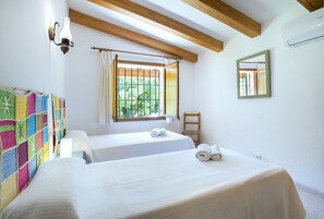 A room with two beds, a colorful quilt, a window with curtains, a wooden ceiling, and a mirror on the wall.
