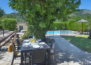 An outdoor dining area with a table set for four, a pool in the background, and a tree providing shade.