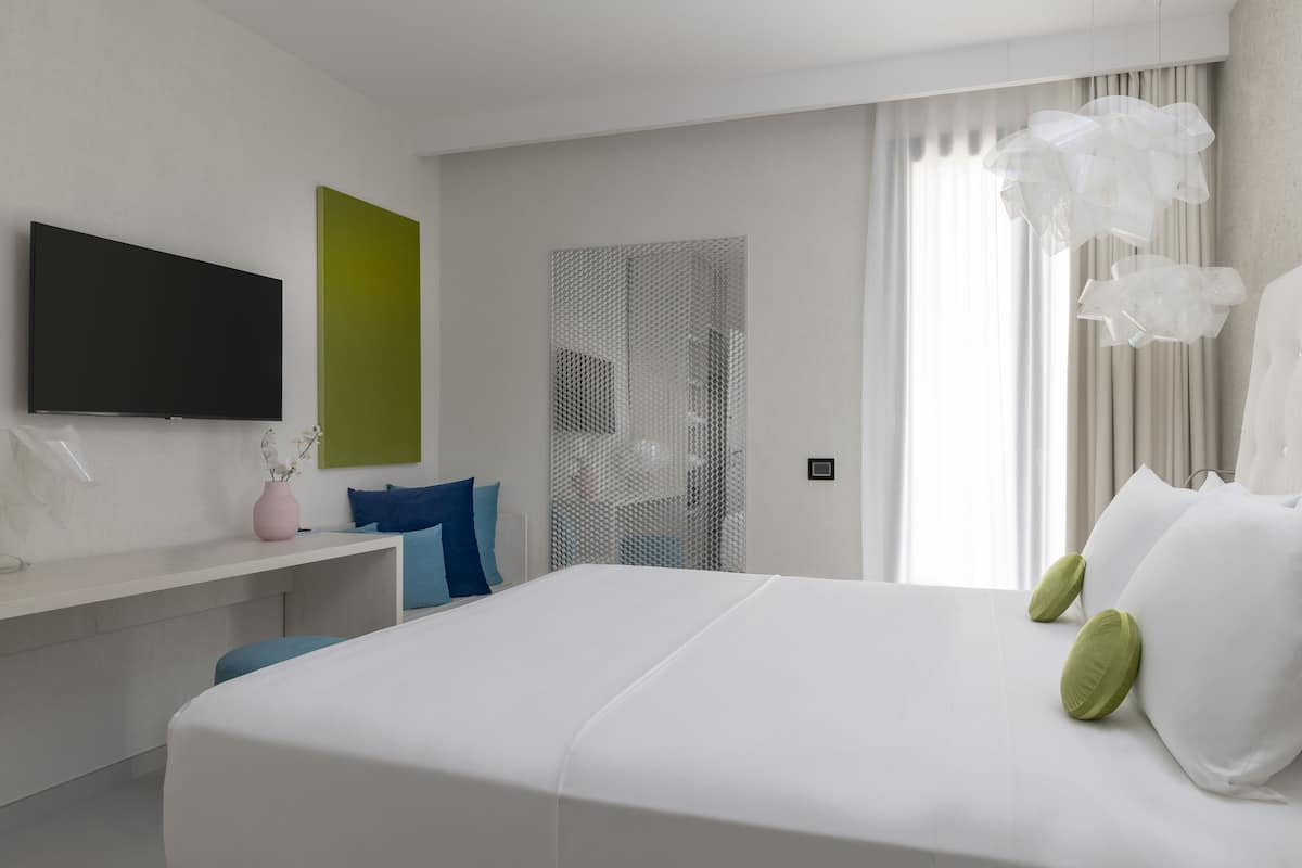 A modern hotel room with a large bed, a flat-screen TV, a desk, and a window with curtains.