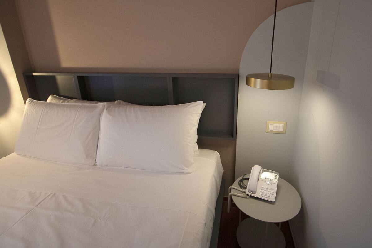 A neatly made bed with white linens, a bedside table with a telephone, and a wall-mounted light fixture.