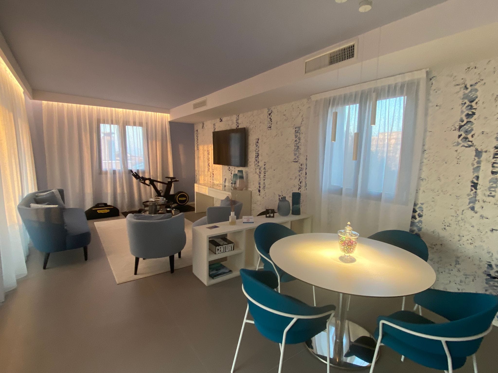 Una sala de estar moderna con una mesa de comedor redonda, sillas azules, un sofá, una bicicleta y un televisor montado en la pared.