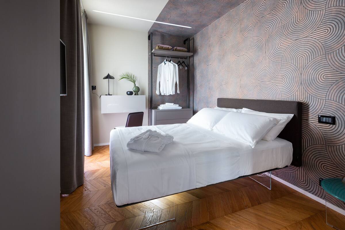 A modern hotel room with a neatly made bed, a desk with a lamp, and a patterned wall.