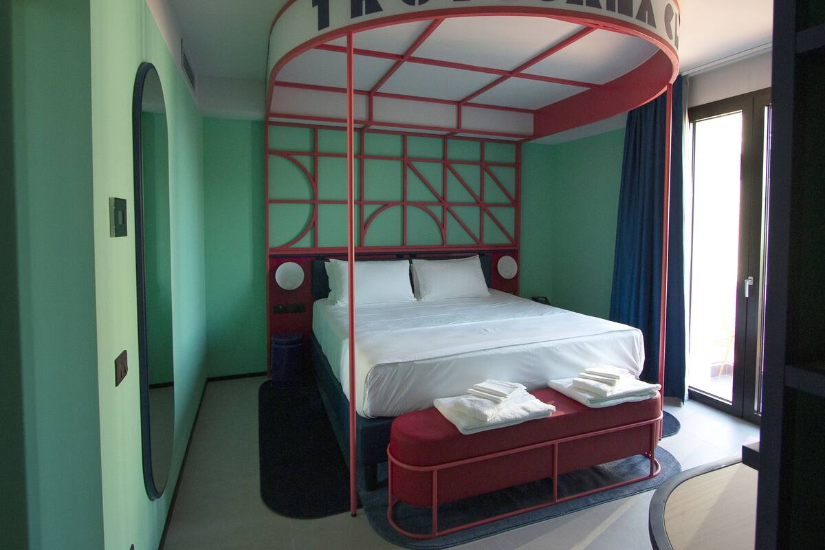 A room with a bed under a red canopy labeled "TROPICANA," a round mirror, and a window with curtains.