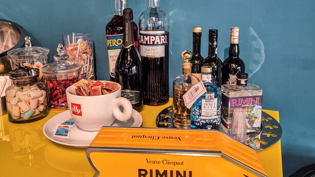 Uma área de bar com diversas garrafas de álcool, uma xícara de café e um tabuleiro com a inscrição 'Veuve Clicquot RIMINI REIMS - 879 KM'.