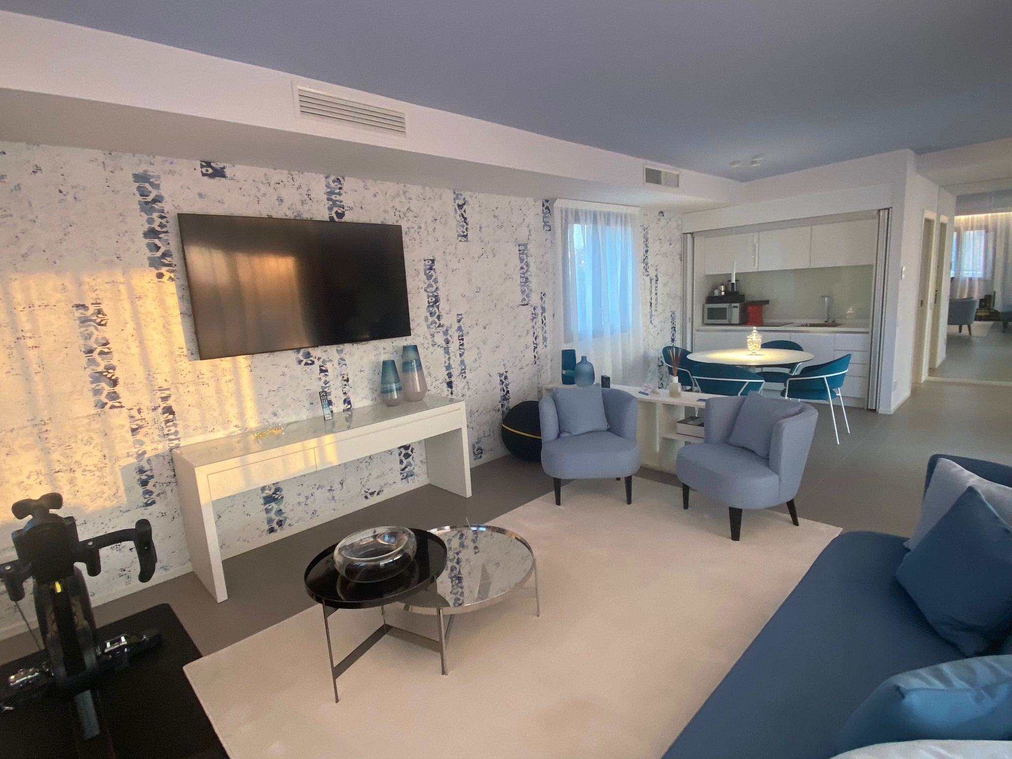 Un salon moderne avec un téléviseur à écran plat, une table basse et un canapé. Au fond, on aperçoit un coin cuisine avec une table à manger et des chaises.