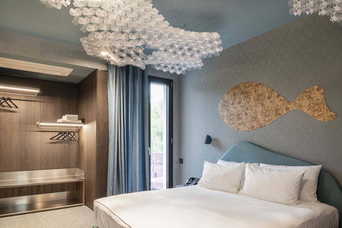 A modern hotel room with a bed, a wall-mounted fish-shaped artwork, a wooden wardrobe, and a large window with curtains.