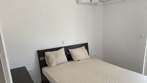 Una cama individual con un cabecero oscuro y dos almohadas, una mesita de noche con un control remoto y un interruptor de luz en la pared.