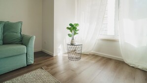 Un salon avec un canapé vert, une petite table d’appoint ronde en fil de fer sur laquelle est posée une plante, et une fenêtre ornée de rideaux légers.