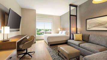 A hotel room with a bed, a sofa, a desk with a chair, a TV, and a window with a view of buildings.