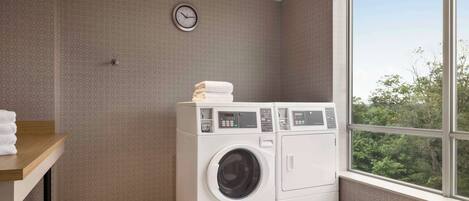 A laundry room with a washing machine and dryer, a clock on the wall, and a window with a view of trees.