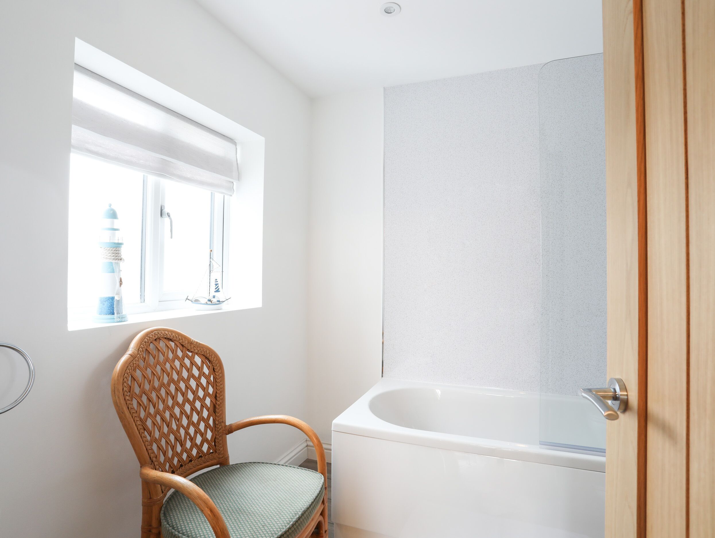 A bathroom with a bathtub, a wicker chair, a window with a view of a lighthouse and sailboat, and a white shower curtain.