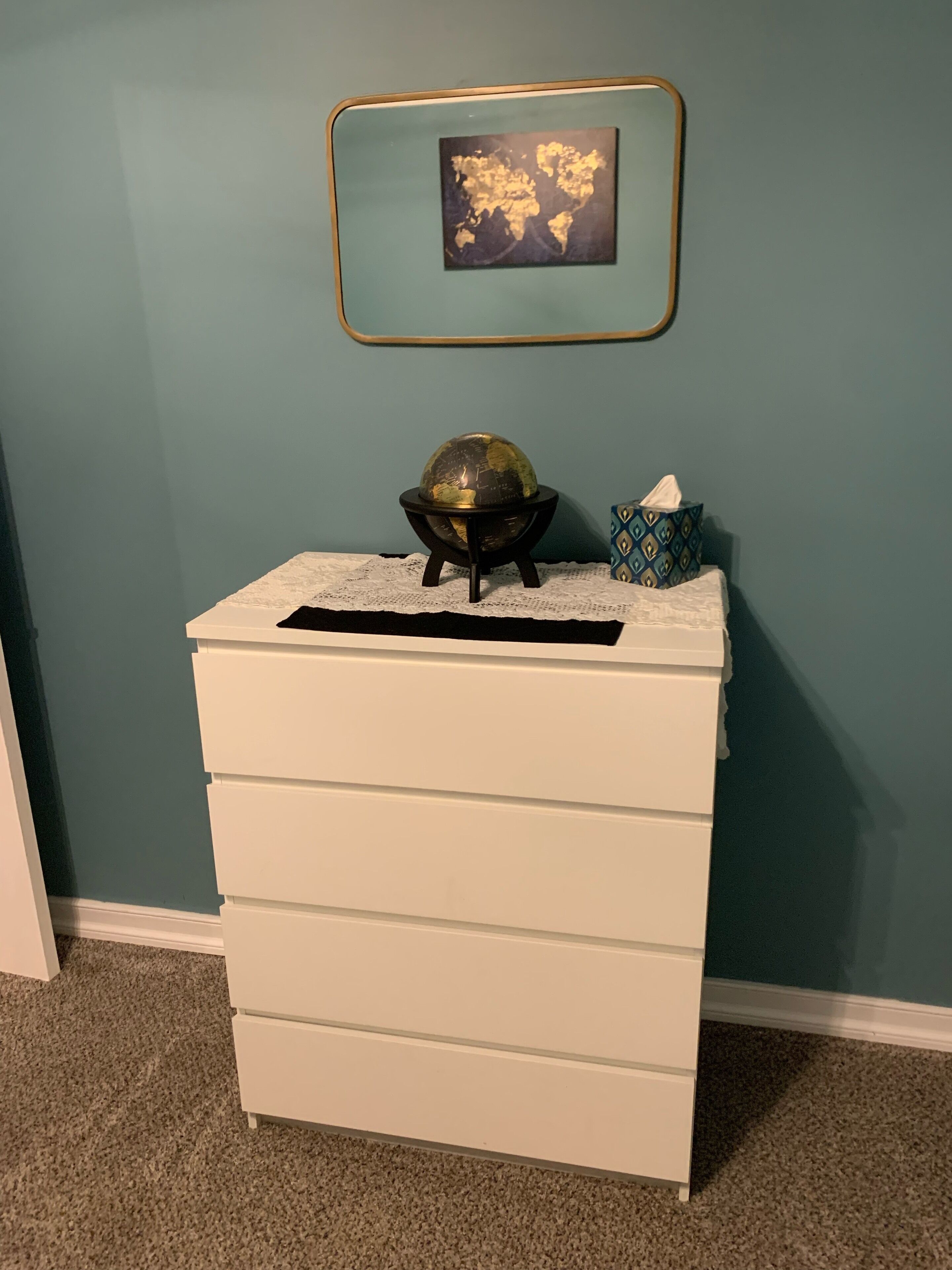 A white dresser with drawers, a globe on top, and a framed picture on the wall.