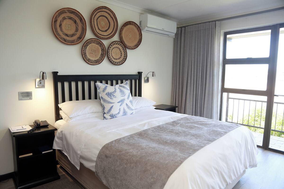 A neatly made bed with a grey blanket, a black nightstand with a phone and a notepad, and a wall with four woven placemats.