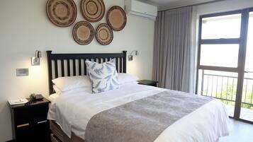 Una cama bien tendida con un edredón gris, una mesita de noche negra con un teléfono y una libreta, y una pared decorada con cuatro individuales tejidos.