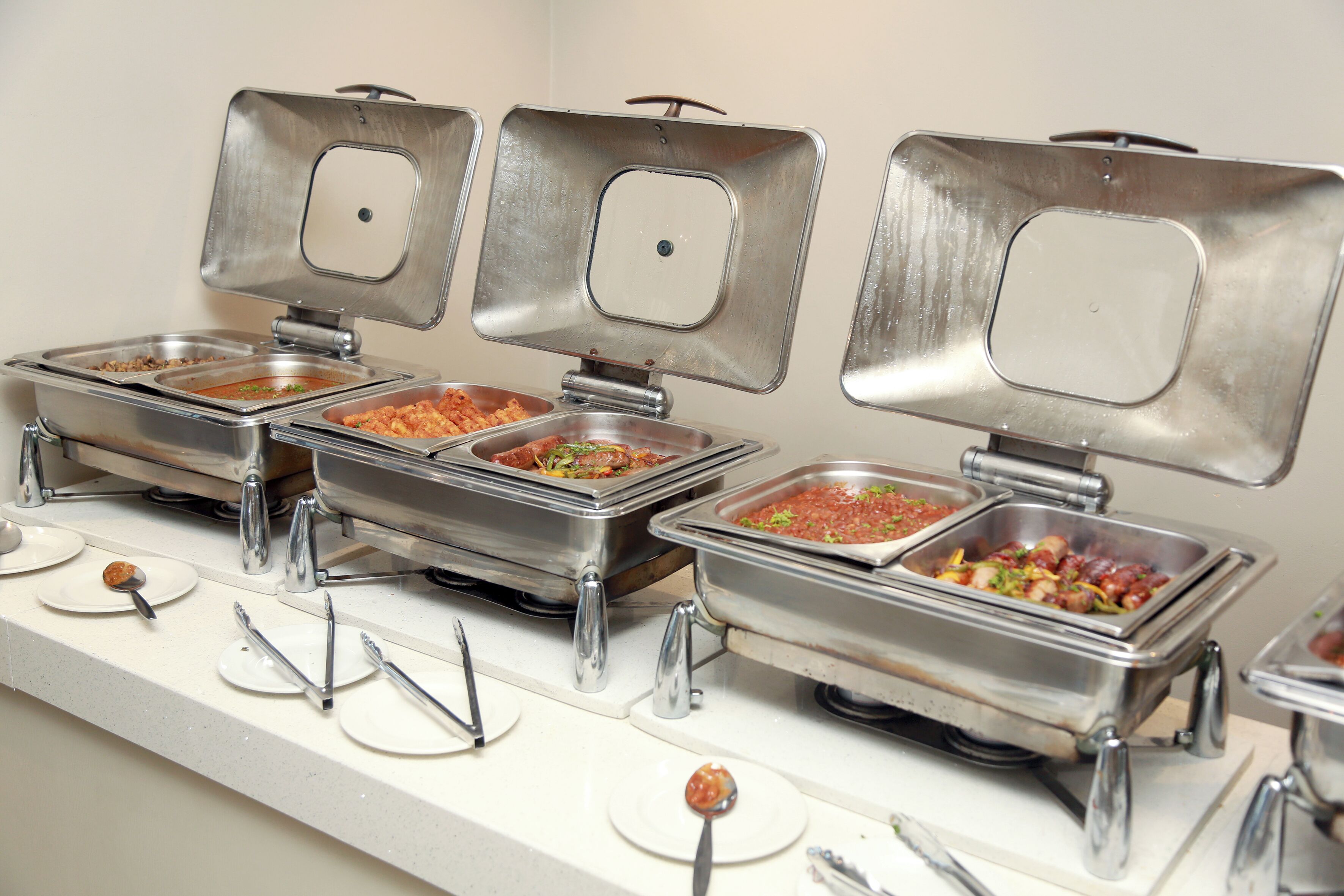 A buffet setup with multiple stainless steel chafing dishes containing various foods, each with a ladle and spoon for serving.