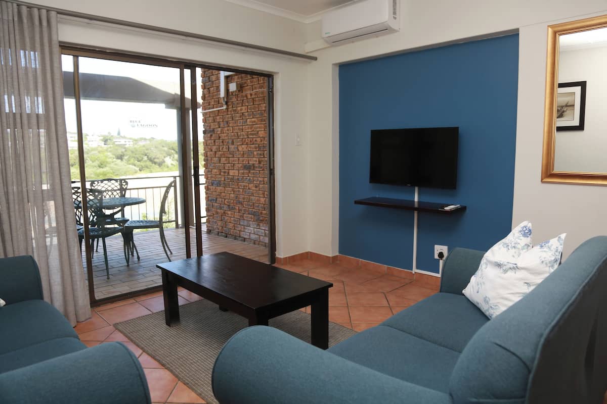 A living room with a sofa, a coffee table, and a TV mounted on the wall. There is a sliding glass door leading to an outdoor area with a table and chairs.