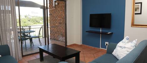 A living room with a sofa, a coffee table, and a TV mounted on the wall. There is a sliding glass door leading to an outdoor area with a table and chairs.