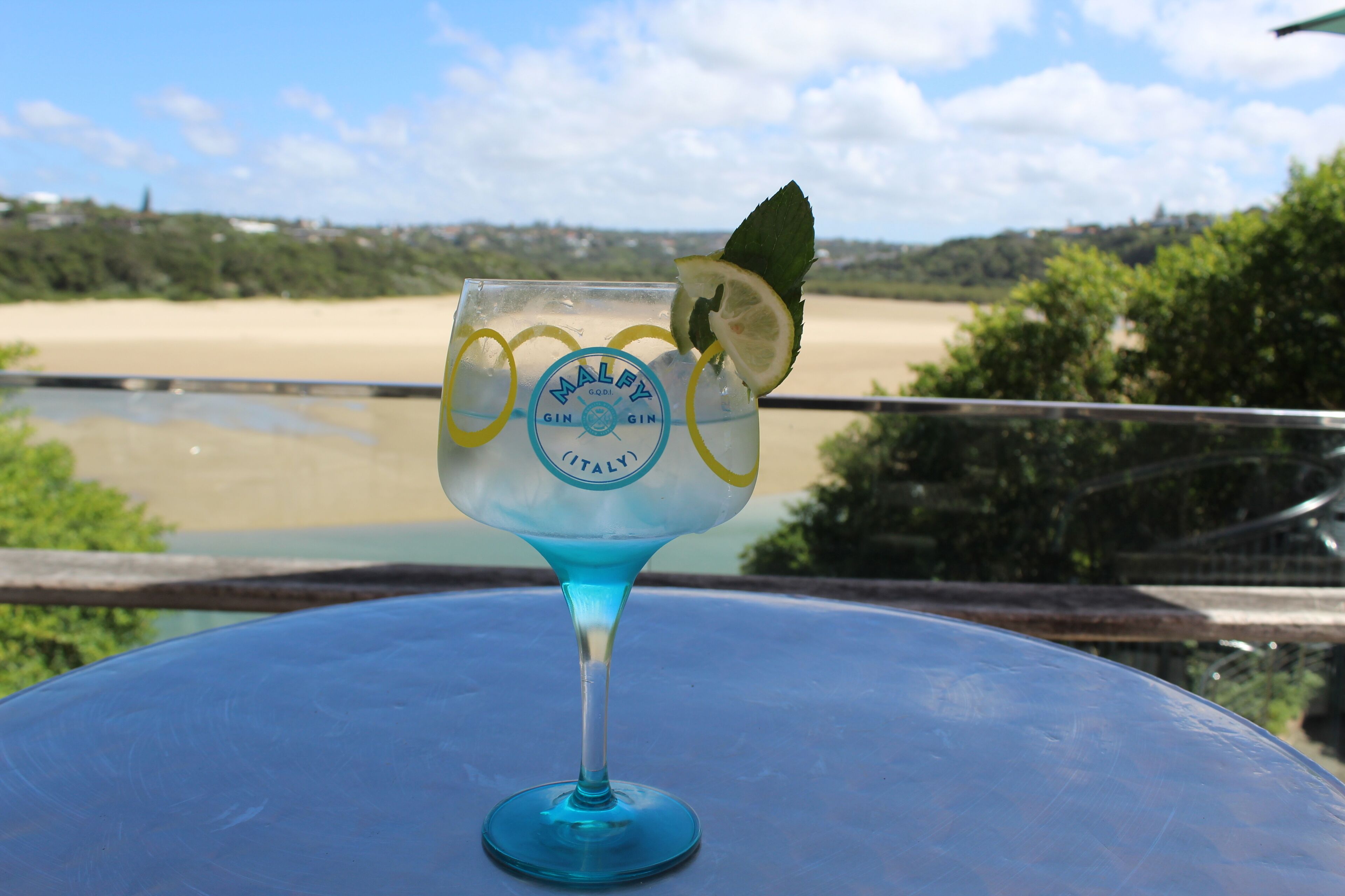 Un verre de cocktail Malfy Gin, garni d’une tranche de citron vert, est posé sur une table d’où l’on aperçoit une plage et des collines en arrière-plan.