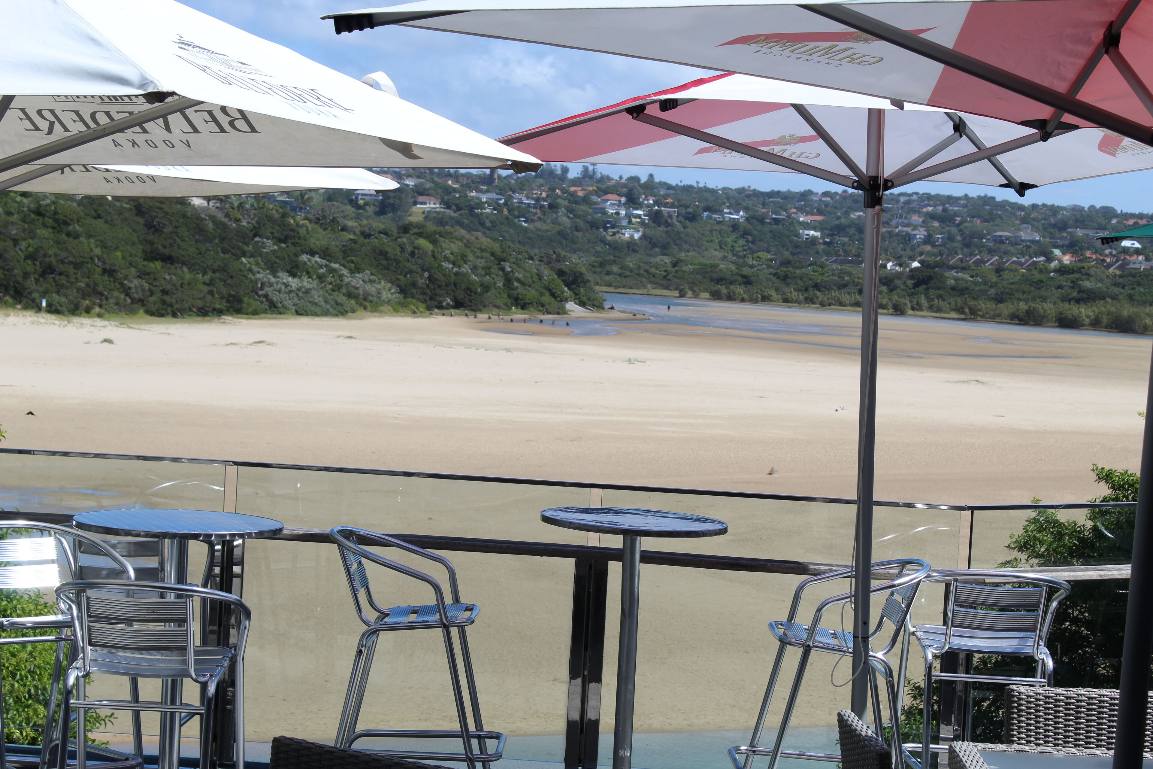 A terrace with metal chairs and tables, overlooking a beach and a hillside.