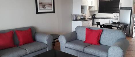 A modern living room with grey sofas and red cushions, a black coffee table, and a kitchen area in the background.